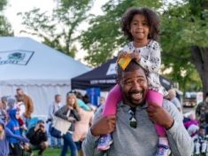 Smiling black daughter sits on smiling black dad's shoulders at a community festival