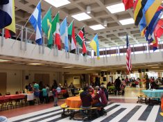 A variety of world flags hang in an atrium where community members mingle and sit at tables