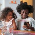 Black woman with a small girl in her lap holds a cellphone and smiles as she looks down at it