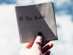 Hand holding a square piece of paper with the words written, #BeKind
