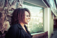 Black woman looks to the right and smiles standing in front of a building with a large window and stone siding